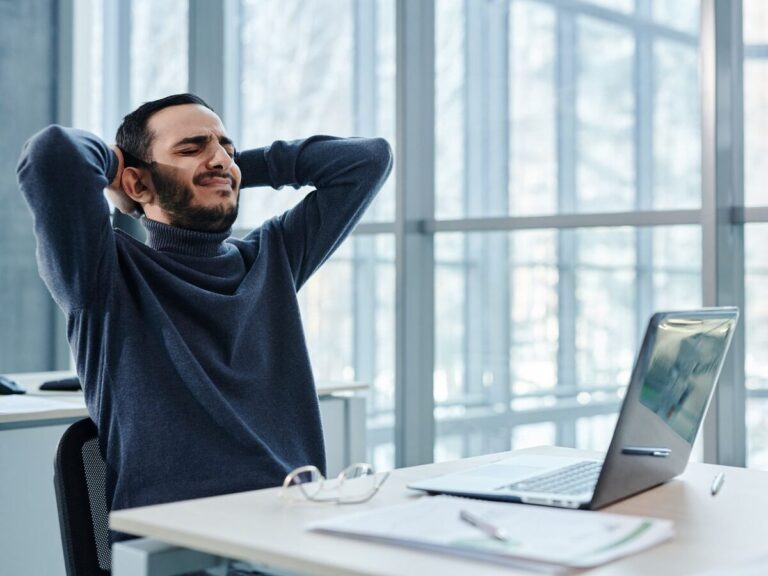 Homme assis à son bureau qui grimace en se tenant la nuque, gêné par une douleur du dos après une longue position assise